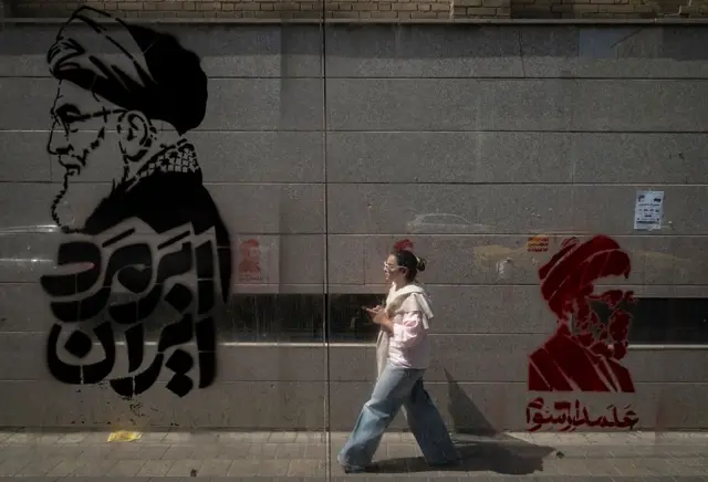 A young Iranian woman walks past graffiti depicting portraits of Iran's late and current Supreme Leaders, Ayatollah Ali Khamenei and Ayatollah Mojtaba Khamenei, in Tehran, Iran, on July 16, 2026.