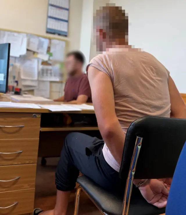 У робочому кабінеті на передньому плані людина сидить на стільці біля письмового столу, тримаючи руки, сковані кайданками, за спиною. Обличчя людей на знімку навмисно розмите. На столі лежать документи та папери, ліворуч видно комп’ютерний монітор і тумбу з шухлядами.