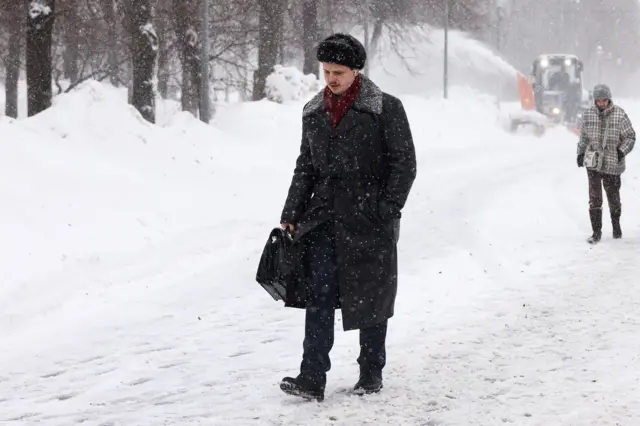 Люди идут мимо высоких сугробов, вдалеке позади едет снегоуборочная машина. Человек впереди одет в пальто и шапку, которые могли бы быть и несколько десятков лет назад, в его руке кожаный портфель. То, что это современная фотография, выдают наушники-капельки у него в ушах.