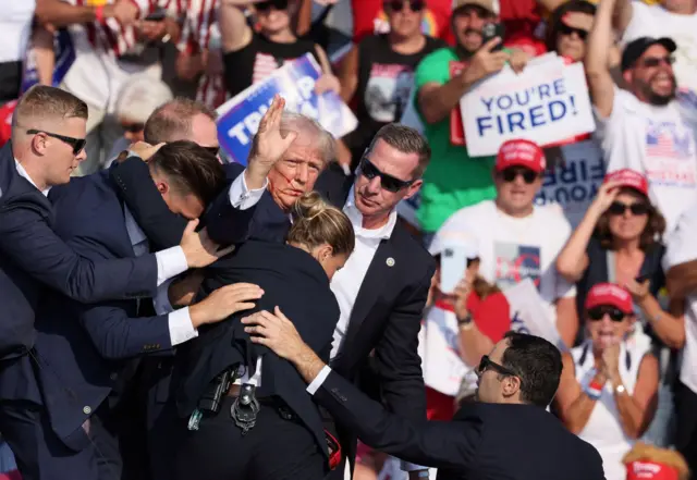 Donald Trump faz gestos com o rosto ensanguentado enquanto é assistido por agentes do Serviço Sereto dos EUA após ser baleado na orelha direita durante um comíciocampanha na Feira AgrícolaButler, na Pensilvânia