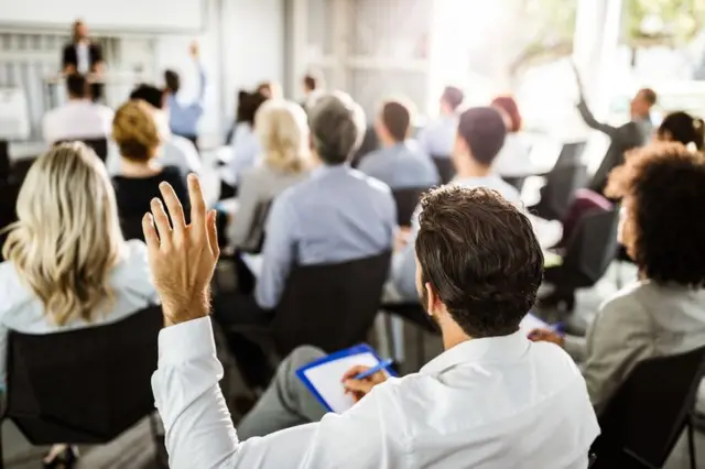 Homem de costas levantando a mão; com outra mão, ele faz anotação. Ele está em sala de reunião com outras pessoas sentadas e outra em pé, liderando o encontro