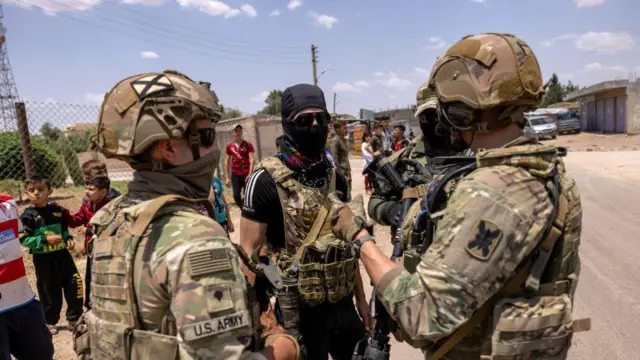 Suriye'nin kuzeydoğusunda, Türkiye sınırına yakın bir bölgede yerel müttefik güçlerle ortak devriye gezen ABD askerleri, bir Kürt askerle konuşuyor. İki askerin arkası dönük, konuştukları kamuflaj yelekli ve silahlı kişi başını ve yüzünü kapatan siyah bere giyinmiş gözlerinde güneş gözlükleri var. Geri planda çocuklar ve siviller var. 