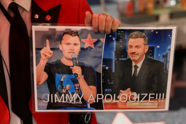 Jimmy Kimmel ve Charlie Kirk'ün yan yana fotoğraflarını gösteren bir döviz. Üstünde "Jimmy özür dile" yazıyor.