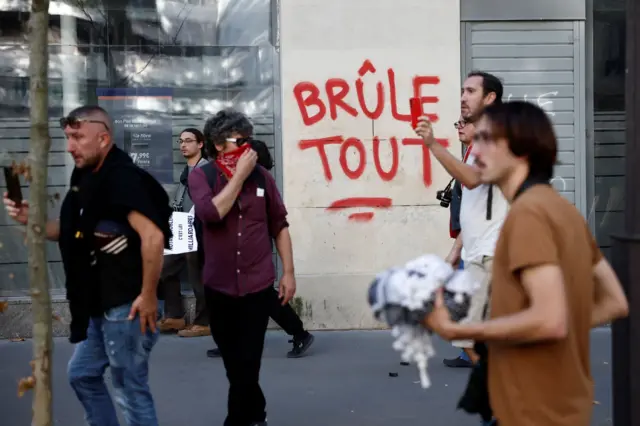 Manifestantes passam diante do slogan “Queime tudo” pintado em um muro.