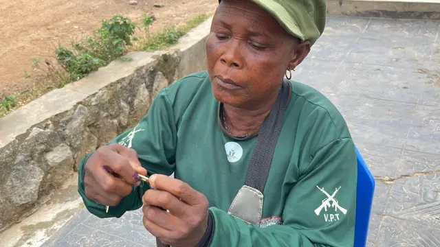 Mama Marian Alarape Iyanuore ree to wọ asọ ologun alawọ ewe, o de fila alawọ ewe pẹlu, o gbe apo alawọ ewe kọ ejika rẹ, o kọ ẹyin si ile kan, to si joko lori aga alawọ buluu, o mu ọta ibọn lọwọ