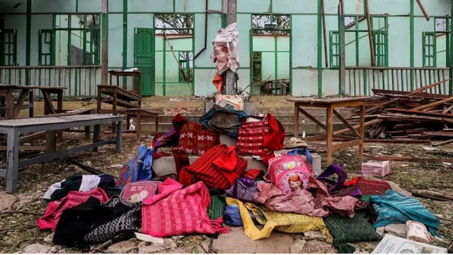 Вещи школьников лежат на земле перед разбомбленным зданием школы
