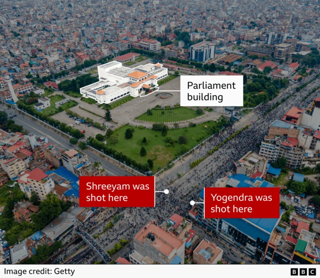 Aerial view of Kathmandu with white parliamentary building set within a green square for di centre. Surrounding di square na dense urban buildings and wide roads. Two red labels mark locations along a road crowded with pipo: one label on di left say “Dem shoot Shreeyam here,” and anoda on di right say “Dem shoot Yogendra here.” One white label above di central complex identify di “Parliament building.” 