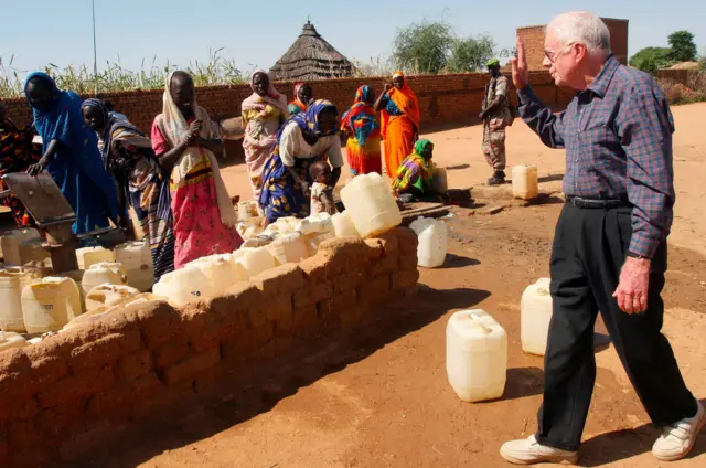 Tsohon Shugaban Amurka Jimmy Carter daga ƙungiyar dattawa, yana ɗaga hannu ga mata ƴan gudun hijira a yayinda suke samun ruwa a birnin Kekkabiya da ke arewacin Darfur a Sudan a 2007.