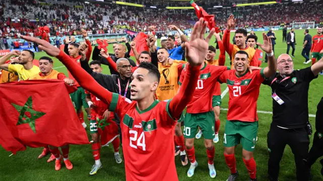 Les joueurs et le staff du Maroc font la fête sur le terrain après avoir battu le Portugal lors de la Coupe du monde 2022 au Qatar. Le groupe s'est organisé selon des lignes approximatives, en plaçant les bras autour des épaules ou en agitant les bras en l'air, avec un joueur, Badr Benoun, séparé au premier plan, près de la caméra. Un autre joueur portant un dossard orange brandit le drapeau du Maroc. Les tribunes d'un stade en train de se vider sont visibles en arrière-plan