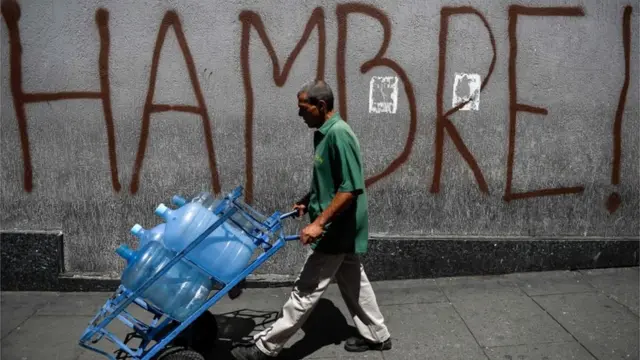 Homem caminha com galões vaziosgalea betágua e a palavra fome está pichada no muro da rua por onde ele passa