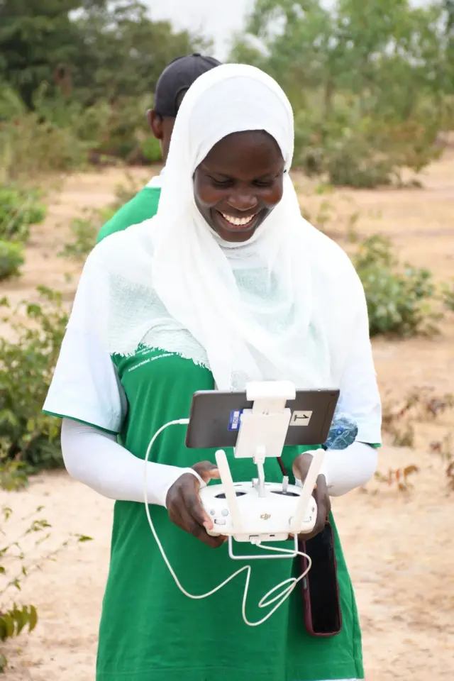 Aminata Sarr porte une robe verte avec une chemise manche longue et un hijab blanc, elle sourit avec dans ses mains un téléphone portable fixé sur une machine blanche.