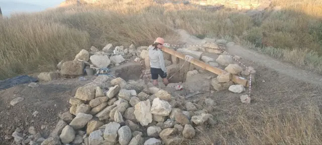 девушка в каске стоит спиной на месте раскопок на фоне груды камней