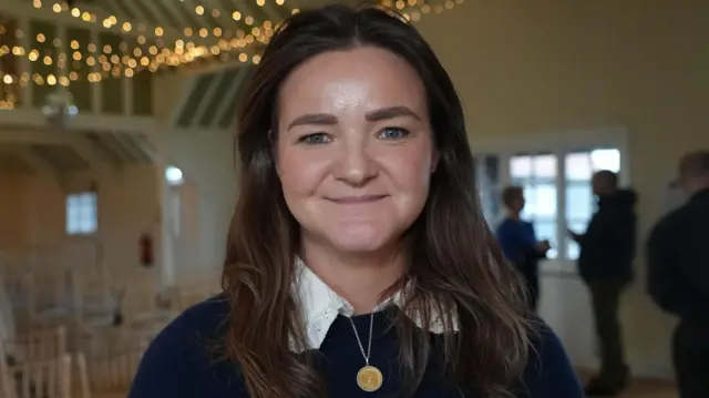 Jenny Riddell-Carpenter, diputada por la costa de Suffolk, mira directamente a la cámara y sonríe. Está de pie en un salón con filas de sillas alineadas detrás de ella. Tiene el pelo castaño y viste una camisa blanca con un jersey oscuro encima y un collar dorado.