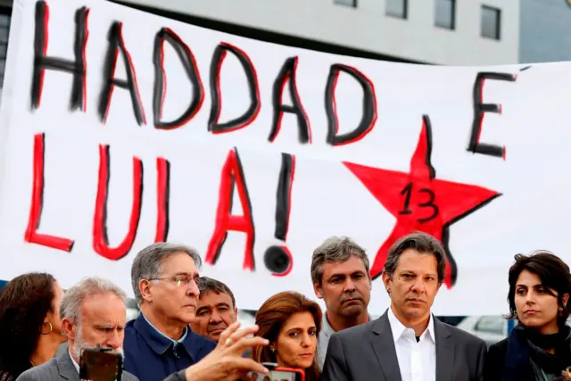 Fotografia colorida mostra uma faixa com a frase "Haddad e Lula! 13" ao fundo e diferentes pessoas da política brasileira no plano à frente, como Fernando Haddad, Manuela d'Ávila, Lindbergh Farias e Ana Estela Haddad