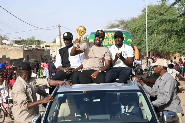 Le sélectionneur national du Sénégal, Pape Thiaw (au milieu sur le toit de la voiture) porte un t-shirt maron avec une casquette noire et tient le trophée dans sa maindroite.