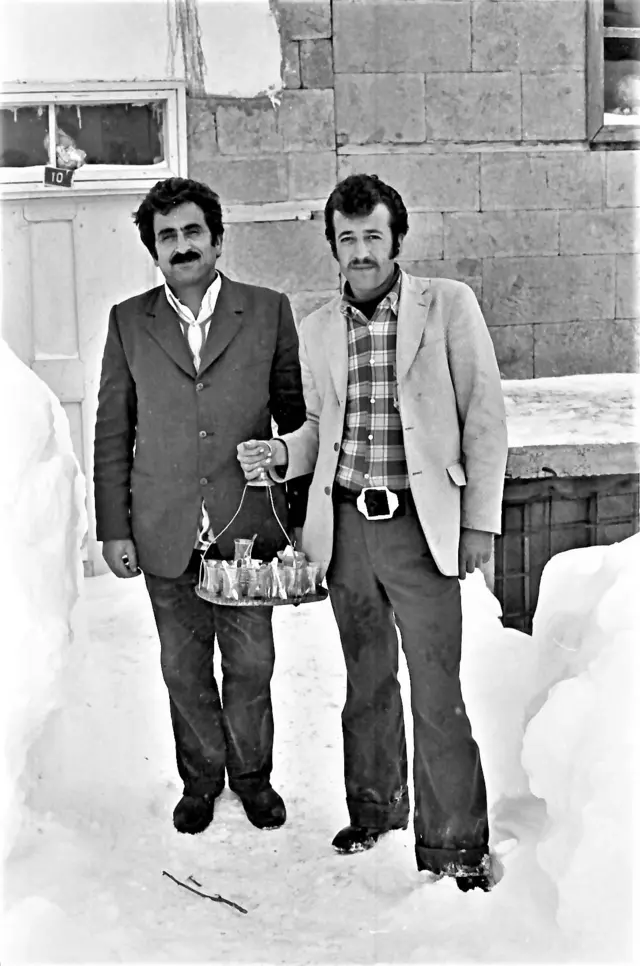 İki adam ayakta, karların yanında poz veriyor. Birinin elinde çay tepsisi var. 