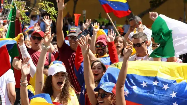 ヴェネズエラ国旗を掲げる人や、国旗色のキャップをかぶった男女たちが笑顔で手をあげて叫んでいる