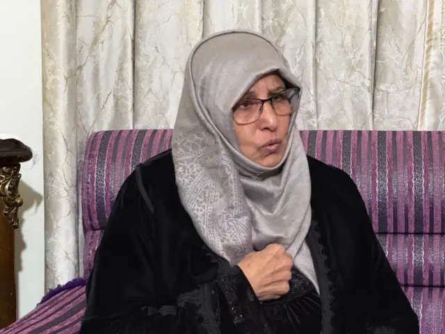 سيدة ترتدي لباساً أسود و حجاباً رمادياً ونظارة طبية تجلس على أريكة بنفسجية اللون خلفها ستارة فضية، تبدو وكأنا تتحدث إلى شخص لا يظهر في الصورة وتبدو عليها علامات الحزن