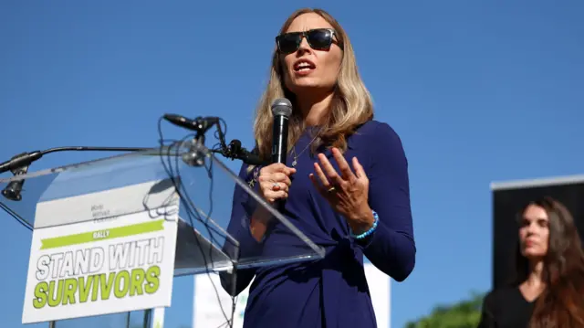 Annie Farmer, elinde mikrofonla, güneş gözlüğü ve resmi mavi bir elbise giymiş halde bir kürsüde duruyor. Hava güneşli ve kürsünün izleyicilere dönük bir panosunda 