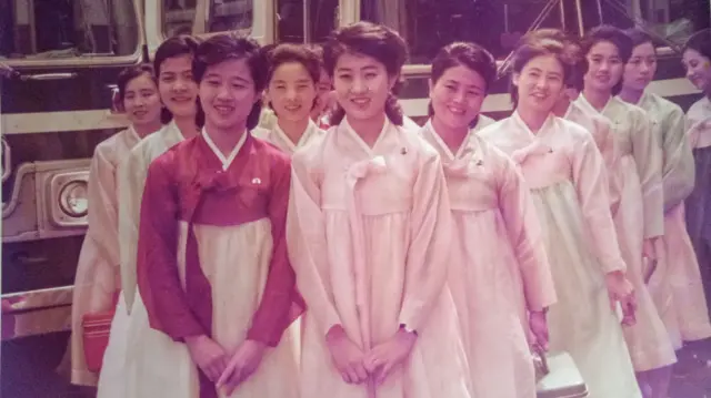 Cerca de uma dúzia de jovens com vestidos tradicionais coreanos estão na frente dos veículos. Ko Yong-hui está na frente, à esquerda 
