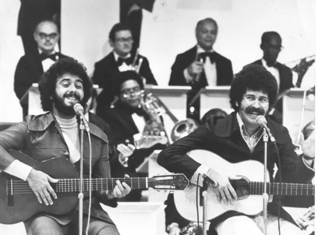 A dupla sentada, cantando, sorrindo e tocando violão em festival, com músicos atrás