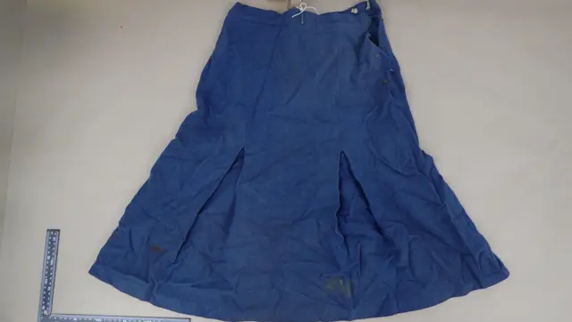 A long blue skirt laid out on a flat surface. There is a section of fabric cut out of the waistband following forensic examination. There is a ruler in the bottom left corner of the frame.