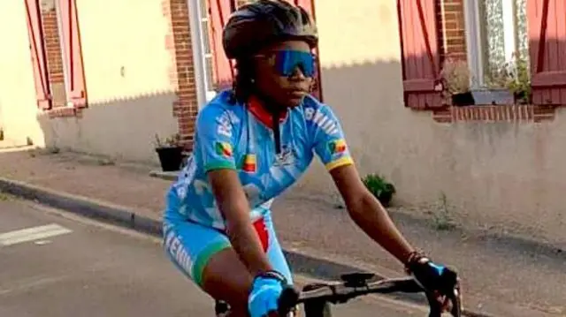 
Georgette Vignonfodo, portant un casque de vélo noir, des lunettes de soleil teintées et des lycras bleu clair, pédale dans une rue.