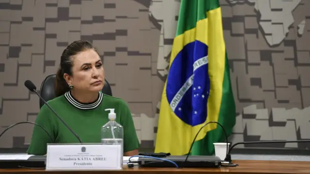 Katia Abreu com olhar sério sentada diantebetboo saque mínimomesabetboo saque mínimocomissão, com bandeira do Brasil ao fundo