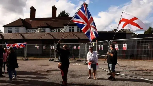 په اېپېنګ سیمه کې د کډوالو د یوه هوټل مخې ته لاریون کوونکي د سېنټ جورج صلیب (ښي اړخ ته) او د یونین جک (کیڼ اړخ ته) بیرغونه پورته کوي