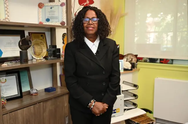 Perpétue Mukarugwiza ahagaze mu cyumba, inyuma ye hari ibihembo mu kabati. Yambaye indorerwamo, ikoti ry'umukara n'ishati y'umweru. Yambaye n'inigi cyangwa ishapule ku kuboko kumwe.