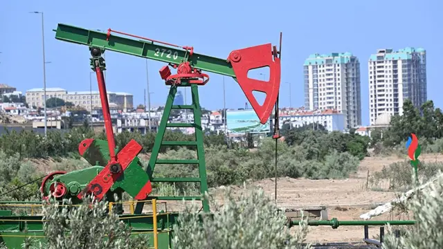 A red and green oil pump at an oil well in the capital city Baku, with apartment blocks, a screen and a park in the background 