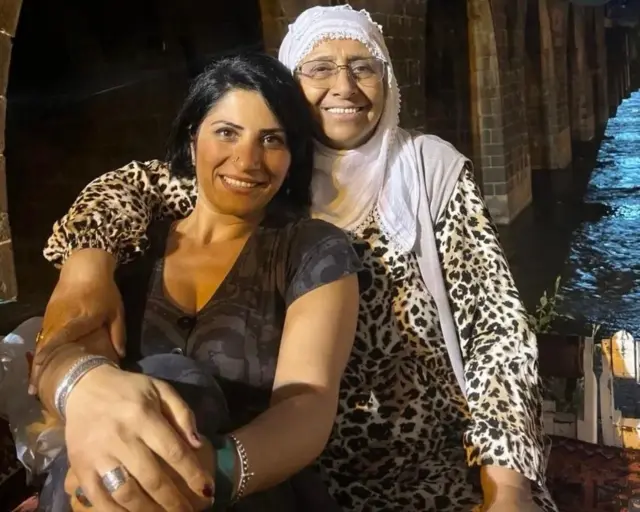 Angel önde; annesi arkada ona sarılmış gülümsüyorlar. 