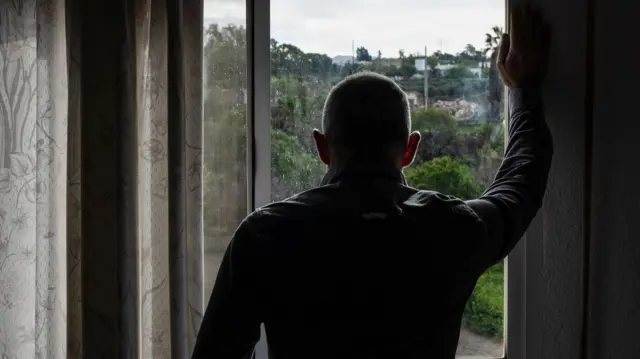 مردی از پنجره به بیرون نگاه میکند، دست راست بالای پنجره تکیه داده و خانهها، درختان و فضای سبز در بیرون.