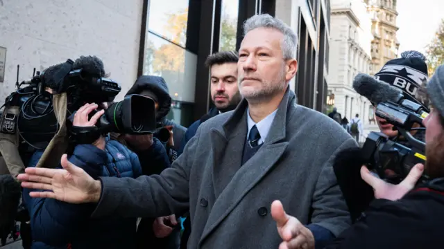 Mężczyzna w szarym płąszczu z wyciągniętymi przed siebie rękami, torujący sobie drogę ludziom trzymającym kamery