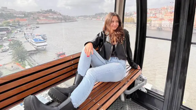 Kaitlin, mặc áo khoác đen và quần jeans, ngồi trong một cabin cáp treo. Phía sau cô là khung cảnh dòng sông trải rộng cùng toàn cảnh thành phố ở phía xa.