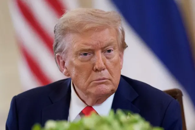 Donald Trump participando de reunião sério, com bandeira dos EUA atrás