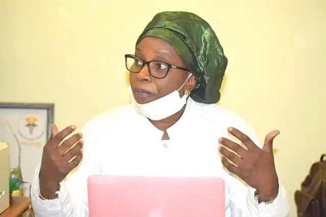 Fatou Samba Ndiaye dans une blouse blanche avec un foulard vert noué sur sa tête, elle parle en levant les deux mains devant un laptop.