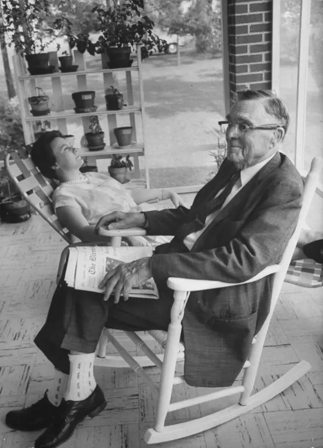 На переднем плане пожилой мужчина в кресле-качалке с газетой в руках, на заднем плане — молодая женщина сидит откинувшись в кресле