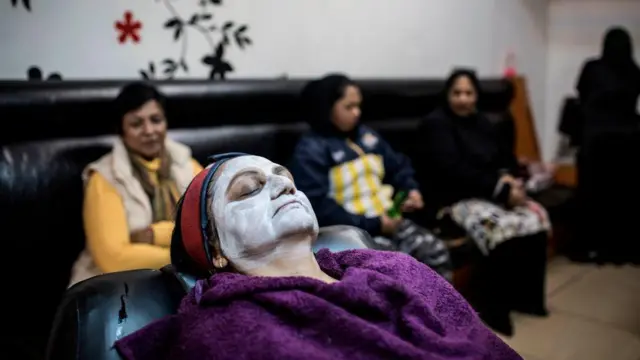 Mulher reclinada com toalha na cabeça e creme para clarear a pele no rosto, com outras mulheres desfocadas ao fundo