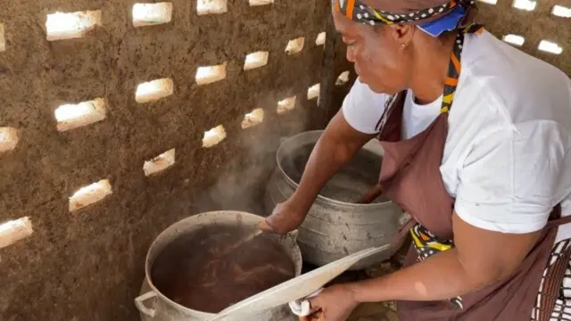 Une femme prépare le beurre de cacao dans une marmite au feu. Elle porte un t-shirt blanc avec un foulard à motif sur la tête.
