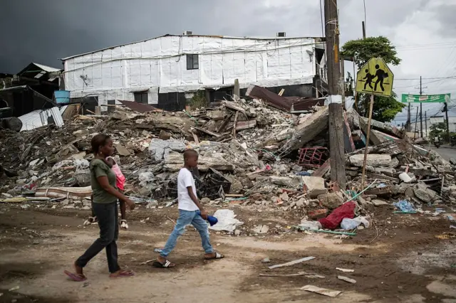 Una mujer camina cargando a su bebé junto a un niño por delante de un edificio en ruinas.