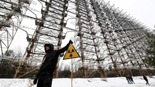 Pessoa de pé em frente a uma enorme antena de radar, ao lado de uma placa com o símbolo de radiação