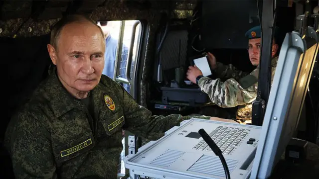 Президент России Владимир Путин в камуфляже внутри военного транспорта сидит перед пультом управления с клавиатурой и экраном, на заднем плане военнослужащий российской армии.