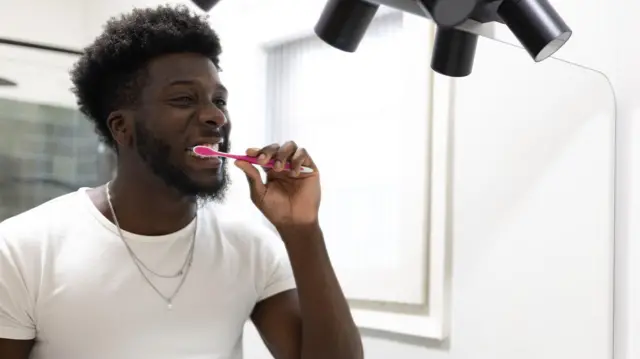 A man looks into the mirror while brushing his teeth. He is wearing a white t shirt and a silver necklace. His toothbrush is pink and the bathroom behind him is white with grey tiles in a shower.