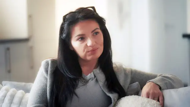 Leanne está sentada em um sofá branco, olhando para alguém com expressão séria. Ela tem cabelos longos e escuros e usa óculos apoiados sobre a cabeça