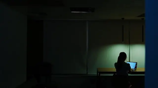 一名女性在昏暗的室內獨自使用筆記型電腦，唯一的燈光照亮她的工作區域。