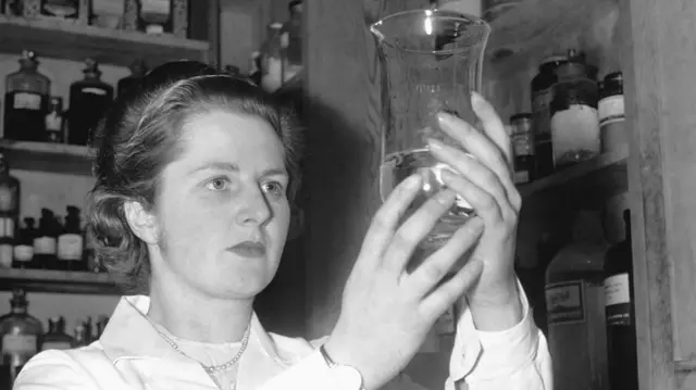 Молодая Юная Маргарет в белом халате рассматривает содержимое стеклянной емкости, на заднем плане видна лаборатория с множеством бутылочек с разной жидкостью на полках
