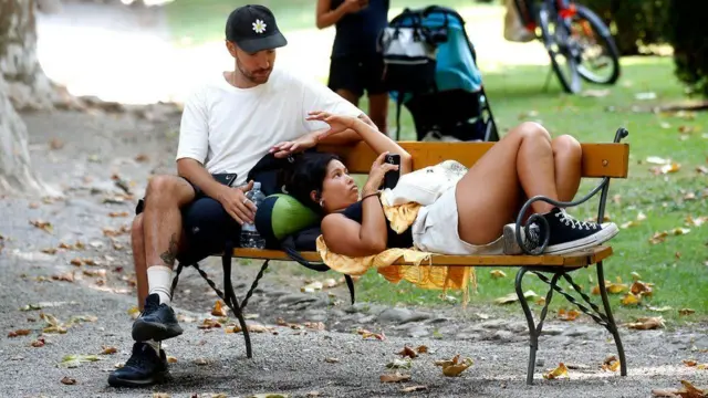 A couple sits on a bench in the park—he’s wearing a white top and navy shorts, while she’s dressed in a black top and white shorts. She is lying on his lap