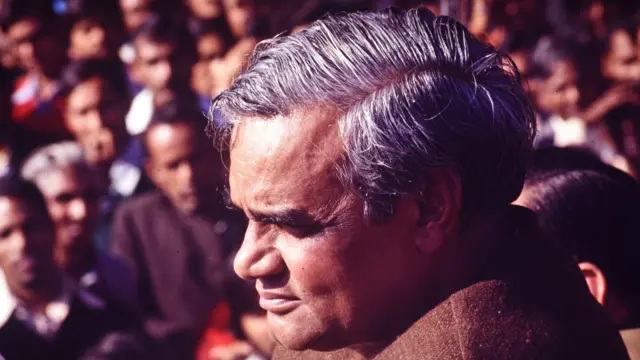 Mężczyzna o siwych włosach, ubrany w brązową kurtkę. W tle widać rozmazany tłum ludzi.