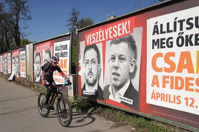 Уздовж вулиці стоїть ряд великих політичних плакатів із портретами Петера Мадяра та Володимира Зеленського. На них написи: «Небезпечні!», «Зупинимо їх!».